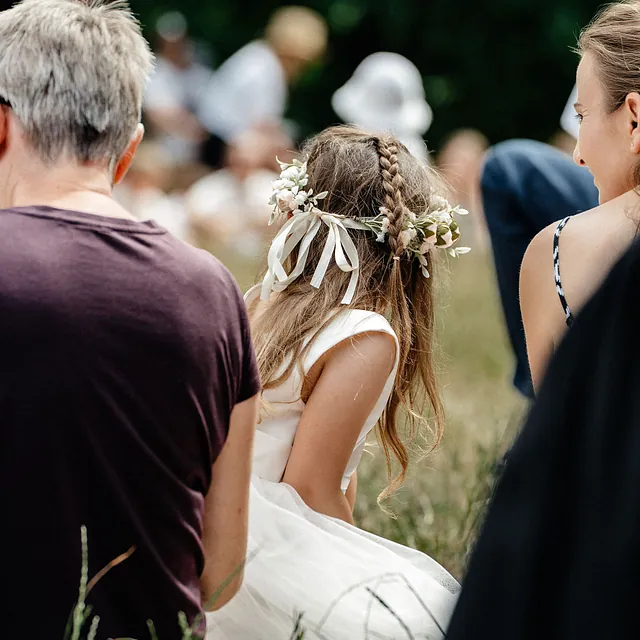 Mädchen mit Blumenschmuck im langen Haar und weißem Kleid von hinten in der Gemeinde (Ausschnitt)