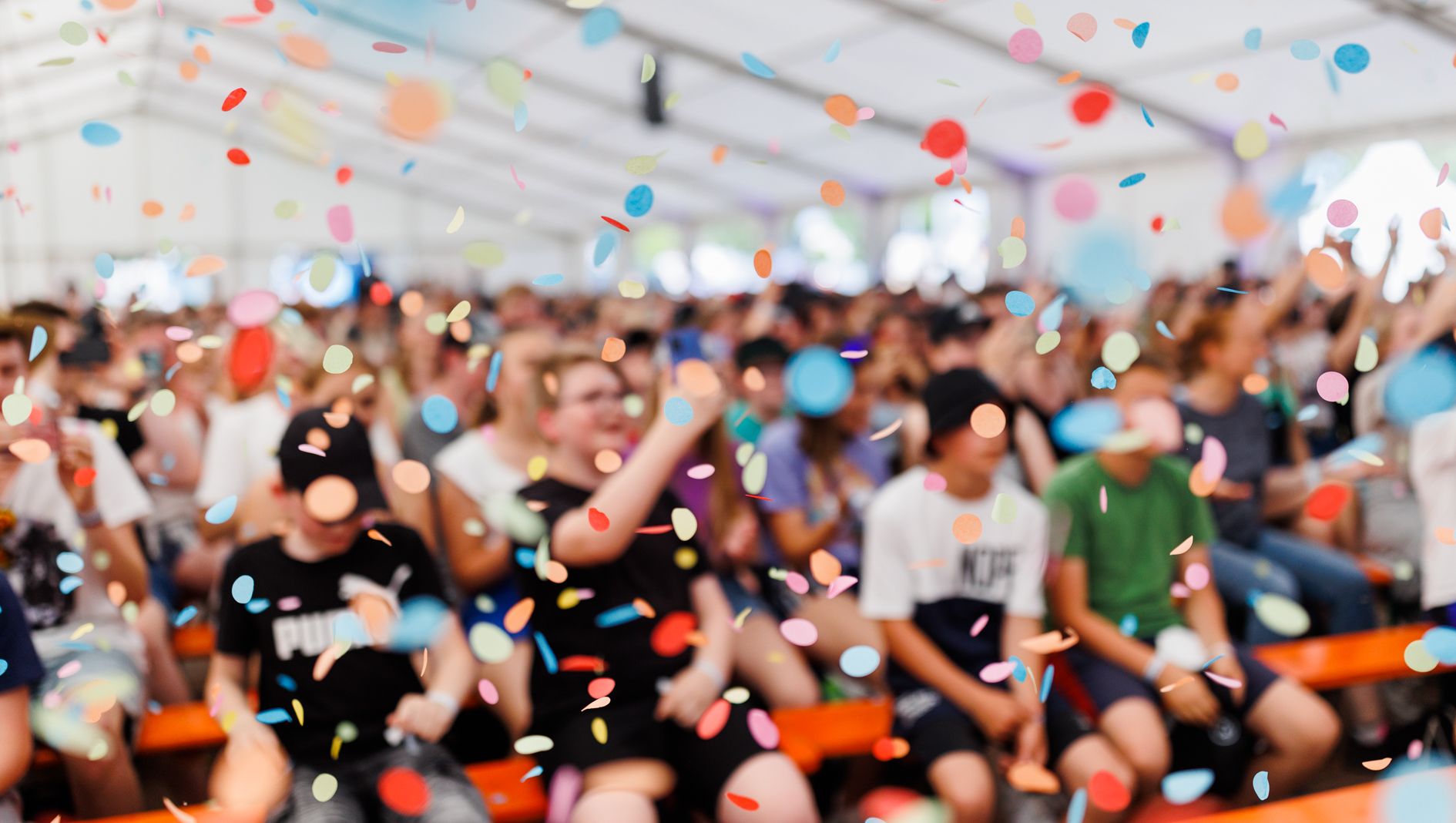 Jugendliche sitzen auf Bänken in einem Zelt, vor ihnen regnet es Konfetti